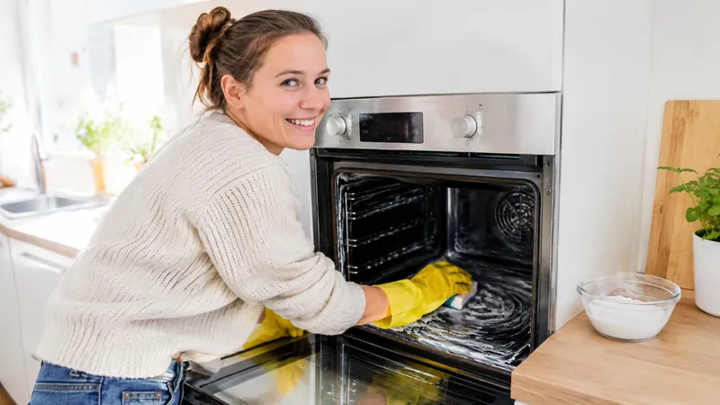 Lächelnde Frau mit gelben Gummihandschuhen streicht weiße Natronpaste in einen offenen Backofen, daneben eine Schüssel mit Paste