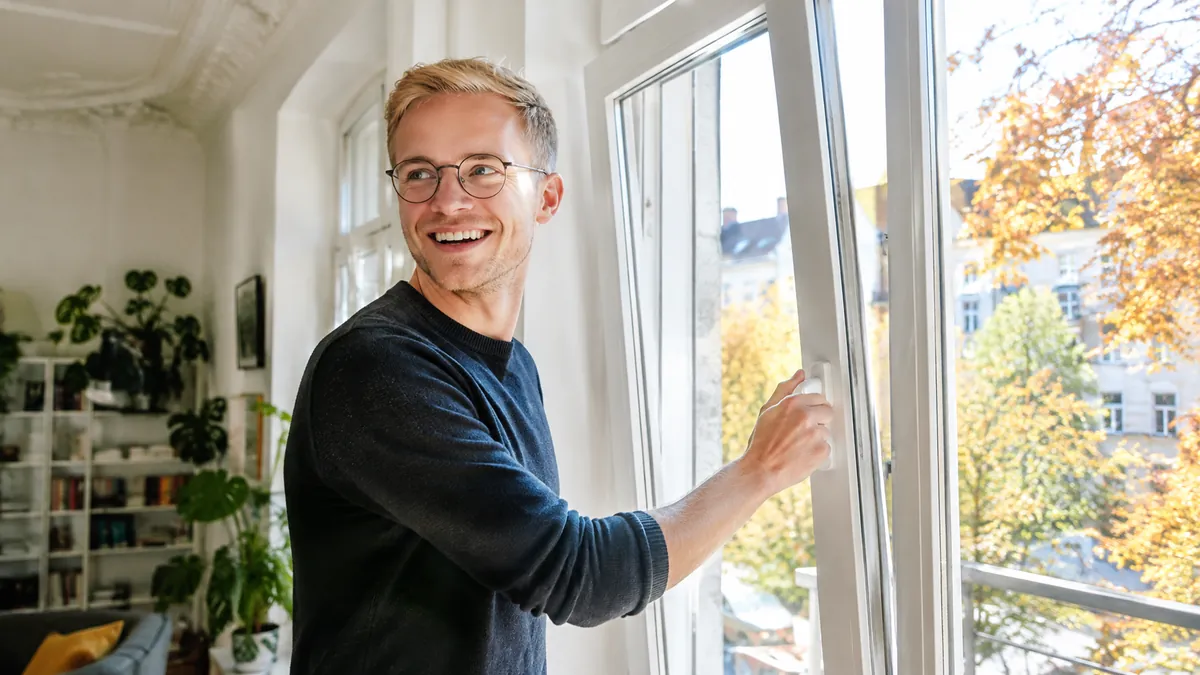 Junger Mann mit Brille hält lachend den Griff eines weißen Dreh-Kipp-Fensters in Kippstellung, heller Altbau, draußen bunte Herbstbäume