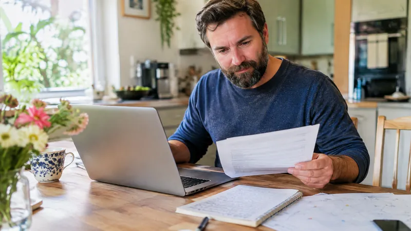Mann in den Vierzigern mit Vollbart vergleicht am Küchentisch Unterlagen auf dem Laptop, daneben Notizblock und Kalender