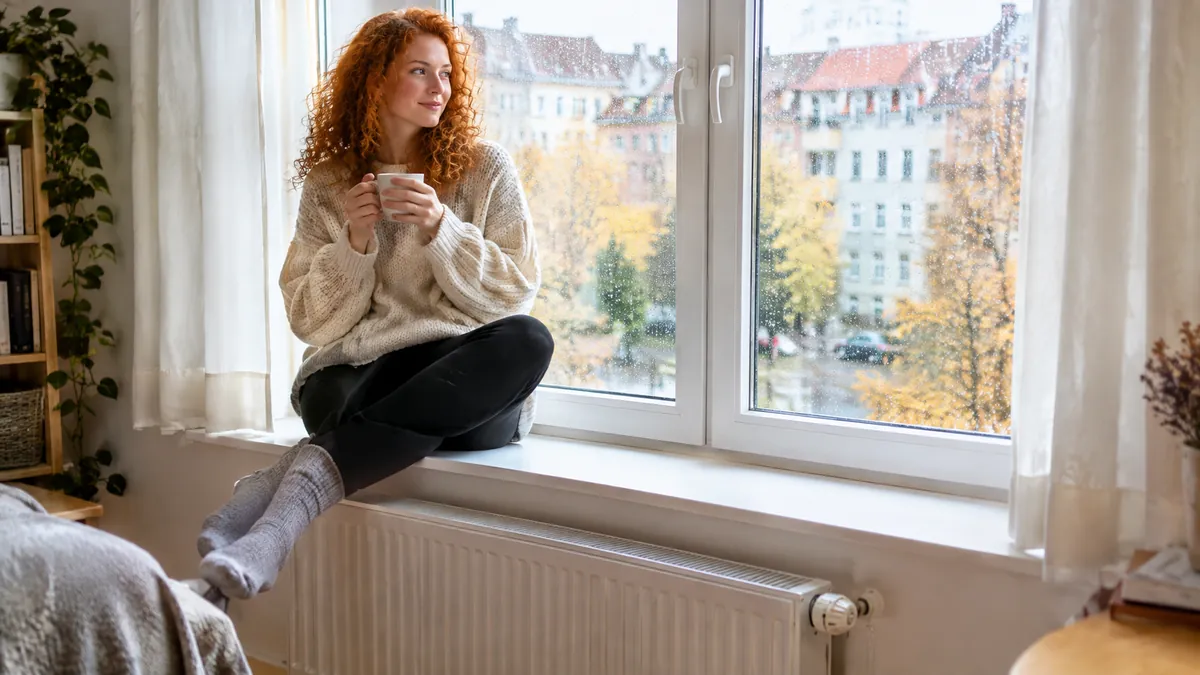 Junge Frau mit roten Locken und Strickpullover sitzt mit einer Tasse auf der breiten Fensterbank, darunter ein weißer Heizkörper, draußen herbstliche Bäume und Altbauten