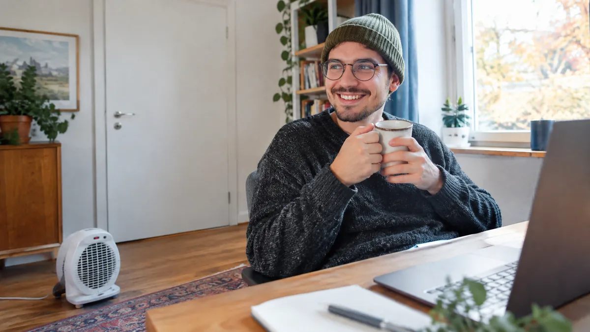 Mann mit Strickmütze und runder Brille lächelt mit Tasse Tee am Schreibtisch, ein weißer Heizlüfter steht mit Abstand auf dem Holzboden