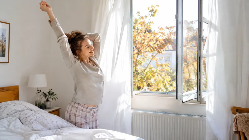 Helles Schlafzimmer mit weit geöffnetem Fenster, weiße Vorhänge wehen, eine Frau streckt sich neben dem Bett