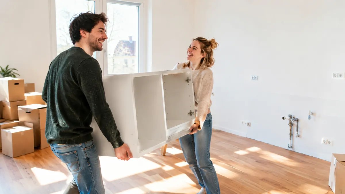 Junges Paar trägt lachend einen weißen Küchenschrank in eine leere Wohnung, an der kahlen Küchenwand sind nur Anschlüsse zu sehen