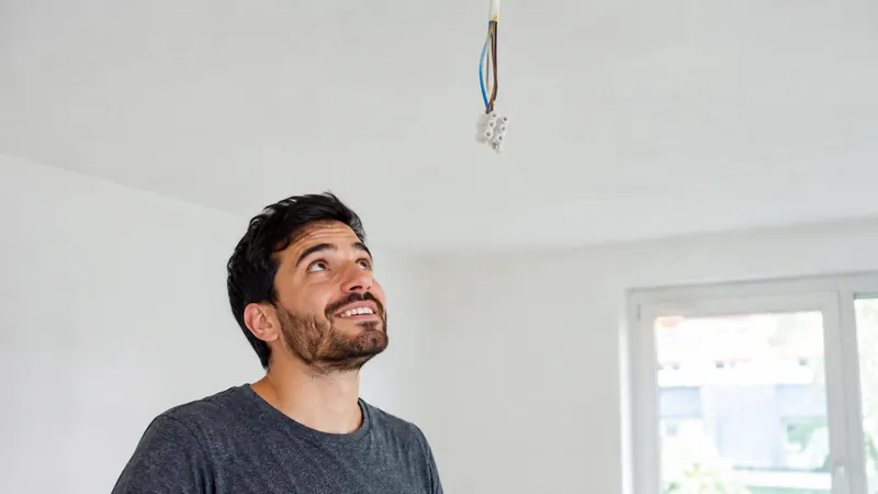 Nackte Kabel einer Deckenlampe hängen von der Decke einer leeren Wohnung, ein junger Mann mit Bart schaut amüsiert nach oben