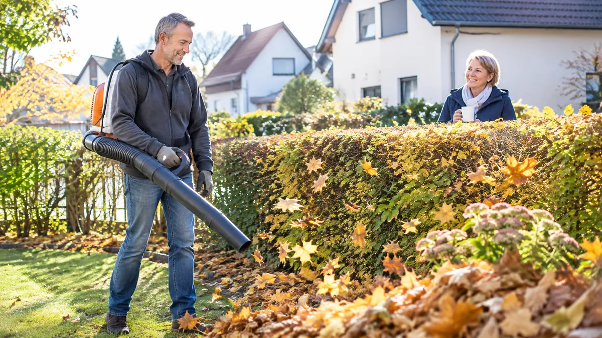 Mann in den Fünfzigern bläst mit einem Laubbläser Herbstlaub im Vorgarten zusammen, hinter der Hecke lächelt ein Nachbar mit Kaffee
