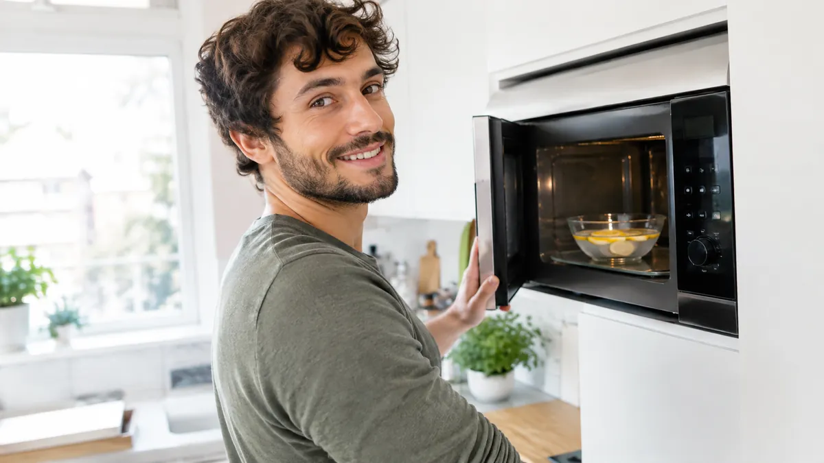 Lächelnder junger Mann mit dunklen Locken und kurzem Bart öffnet in heller Küche eine Mikrowelle mit Glasschüssel und Zitronenscheiben