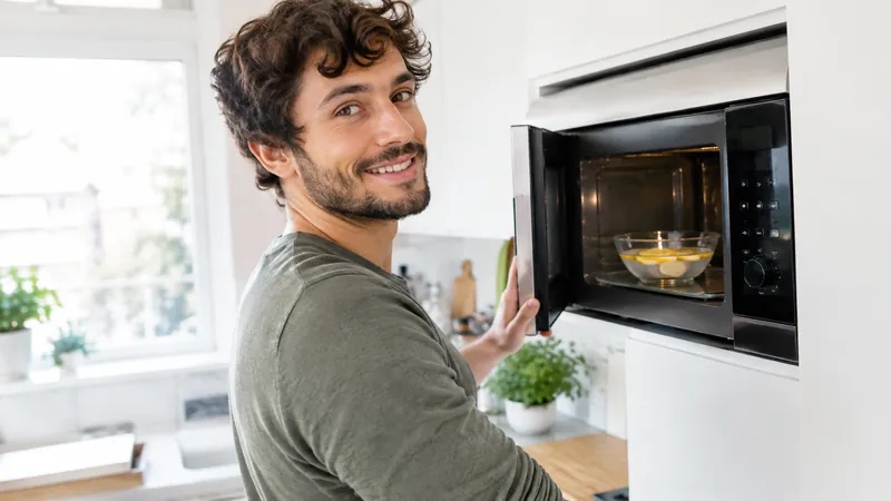 Lächelnder junger Mann mit dunklen Locken und kurzem Bart öffnet in heller Küche eine Mikrowelle mit Glasschüssel und Zitronenscheiben
