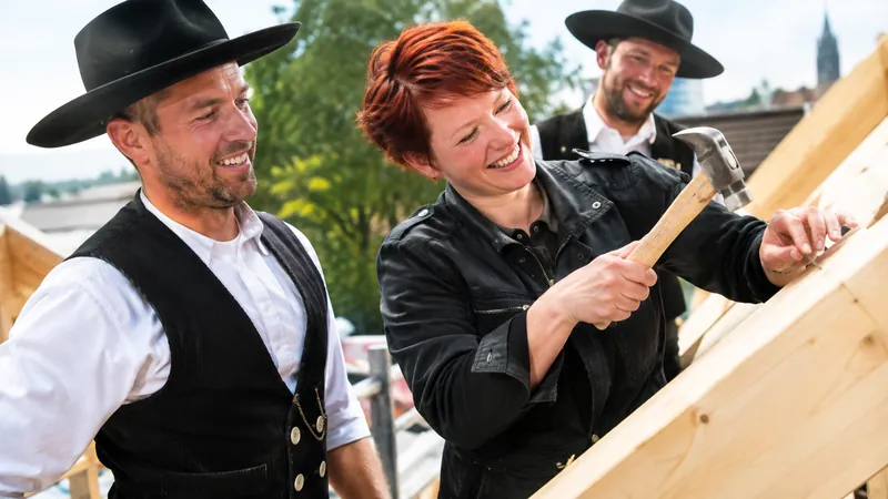 Paar, die Frau mit kurzen roten Haaren, schlägt beim Richtfest einen Nagel in einen Holzbalken, Zimmerleute mit schwarzen Hüten daneben
