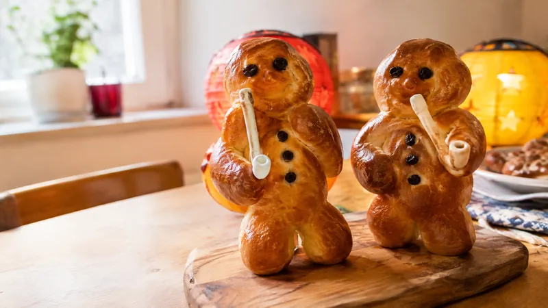 Zwei Weckmänner aus Hefeteig mit Rosinenaugen und kleiner weißer Tonpfeife auf einem Holzbrett, im Hintergrund leuchtende Papierlaternen