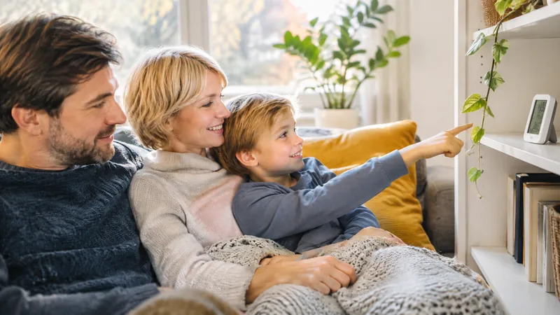 Familie zu dritt unter einer Strickdecke auf dem Sofa, der kleine Sohn zeigt auf ein digitales Raumthermometer im Regal