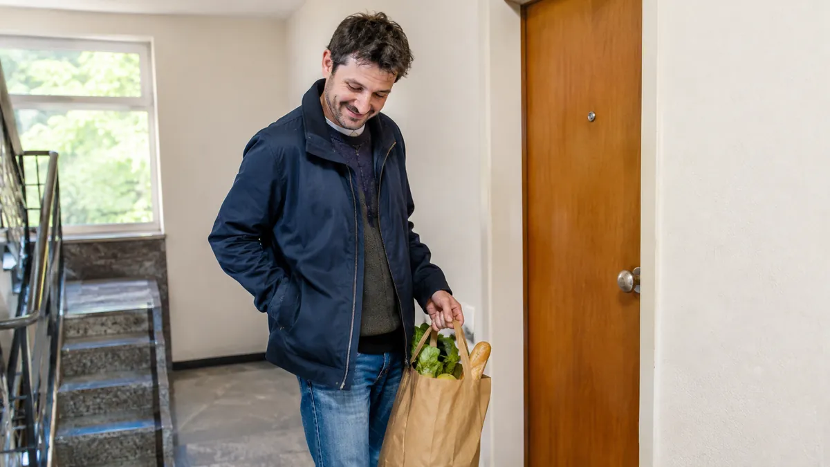 Mann in den Dreißigern mit Einkaufstüte klopft im Treppenhaus vor seiner geschlossenen Wohnungstür mit Knauf lächelnd die Jackentaschen ab