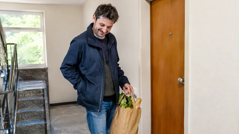 Mann in den Dreißigern mit Einkaufstüte klopft im Treppenhaus vor seiner geschlossenen Wohnungstür mit Knauf lächelnd die Jackentaschen ab
