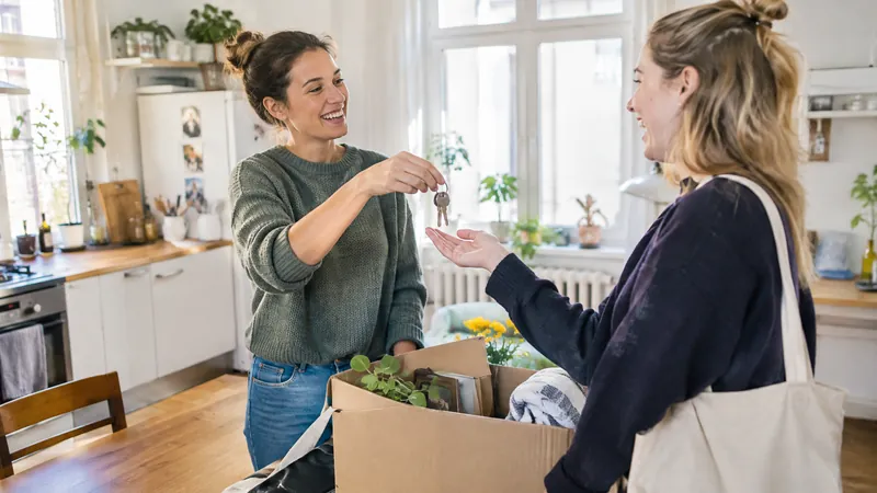 Zwei junge Frauen lachen in einer hellen Altbau-WG-Küche, eine reicht der anderen einen Zimmerschlüssel, auf dem Tisch ein Umzugskarton