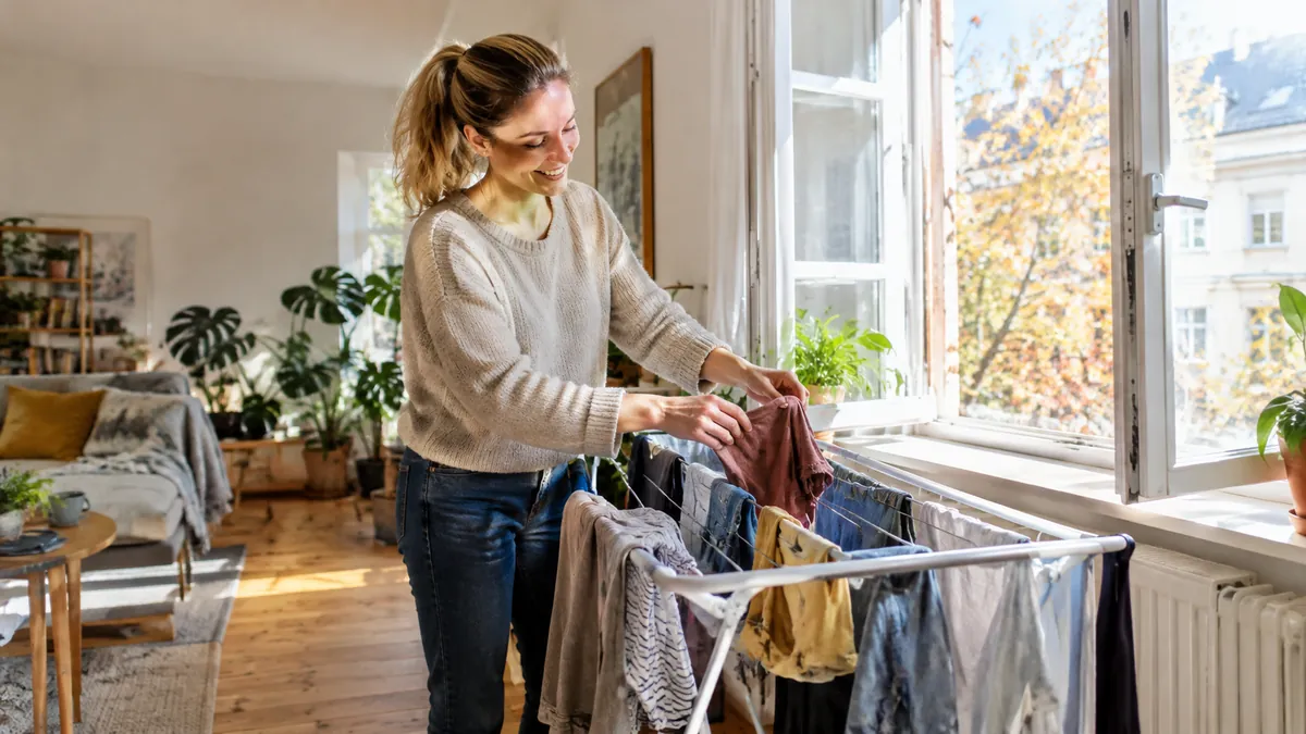 Lächelnde Frau hängt bunte Wäsche auf einen Wäscheständer vor einem weit geöffneten Fenster in einem hellen Altbauzimmer