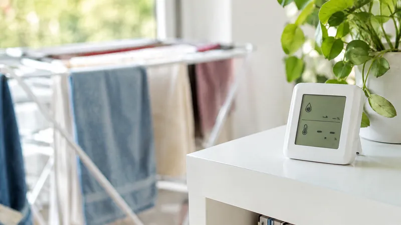 Kleines digitales Hygrometer auf einem weißen Tisch neben einer Grünpflanze, im Hintergrund unscharf ein Wäscheständer mit Handtüchern